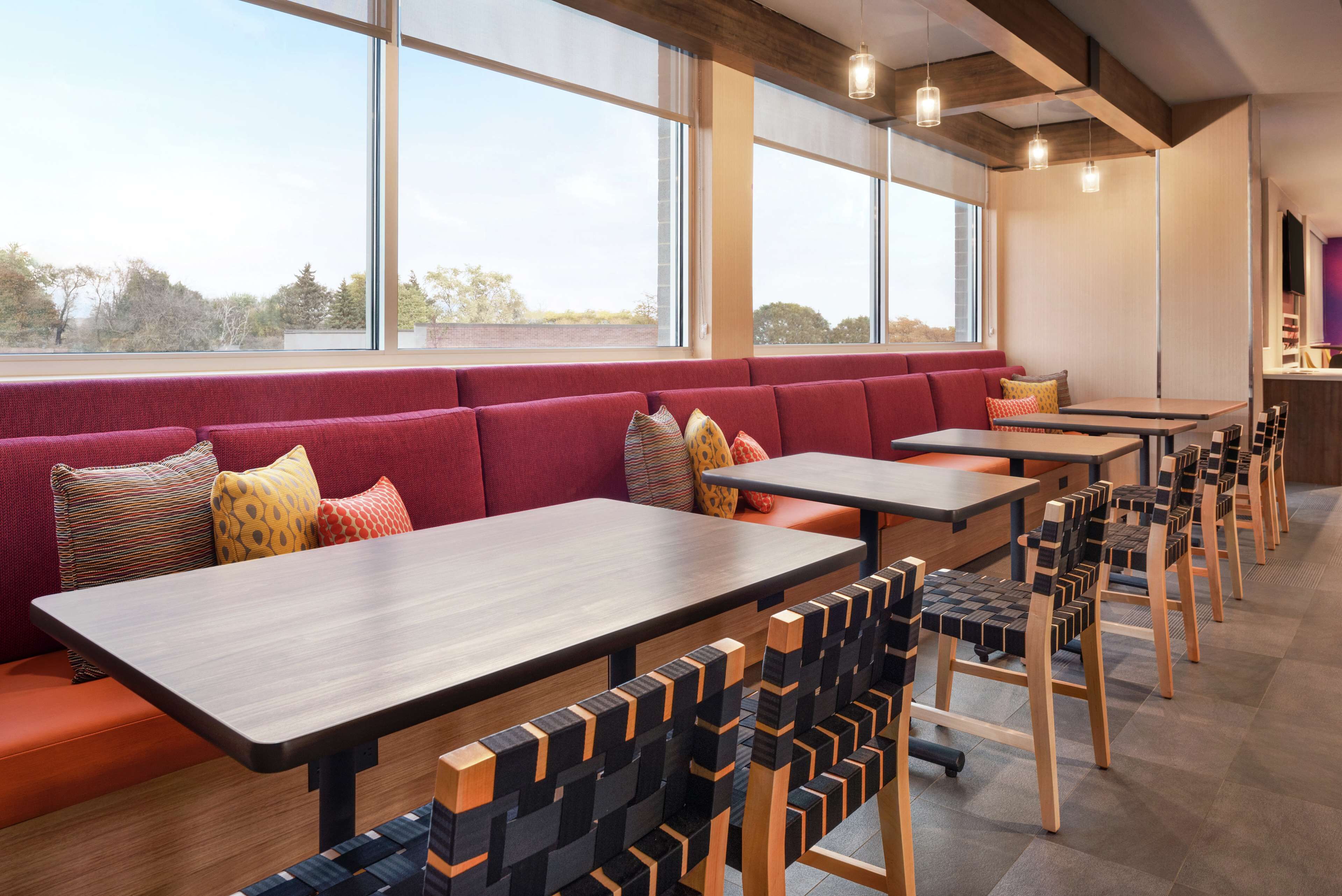 Un espace de restauration moderne avec des tables et des chaises en bois, un banc rembourré de couleur rouge et de grandes fenêtres offrant une vue sur l’extérieur.