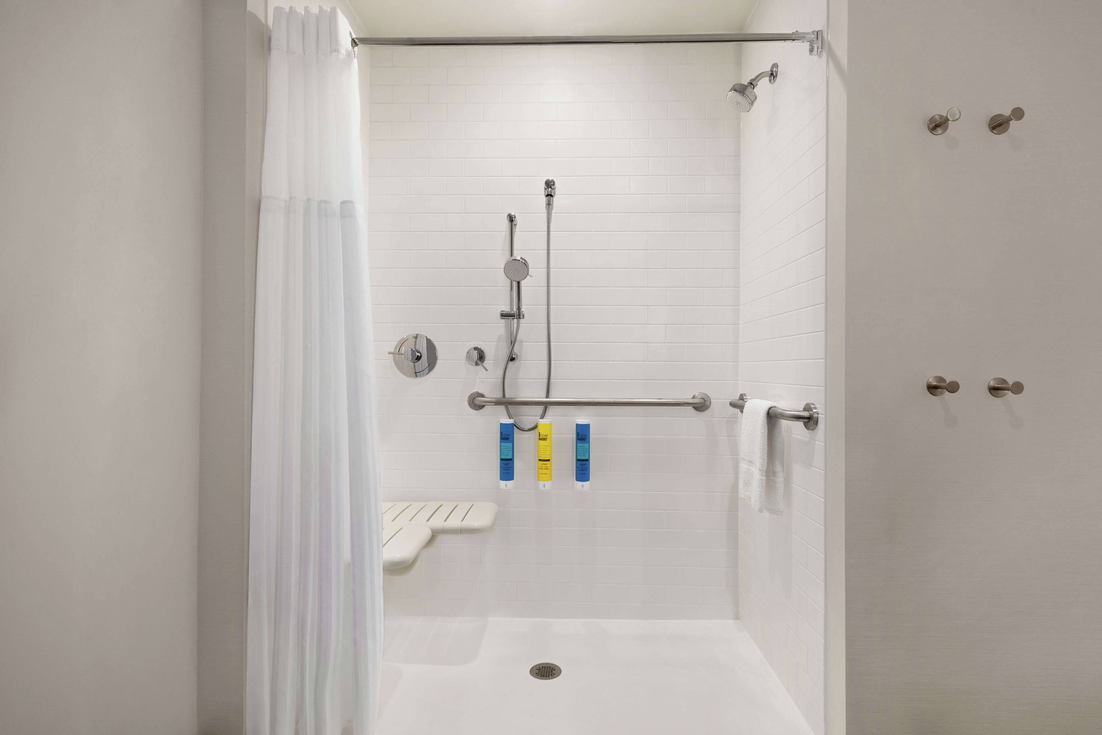 Ducha a nivel con paredes de azulejos blancos, cabezal de ducha, ducha de mano, barra de apoyo y toallero.