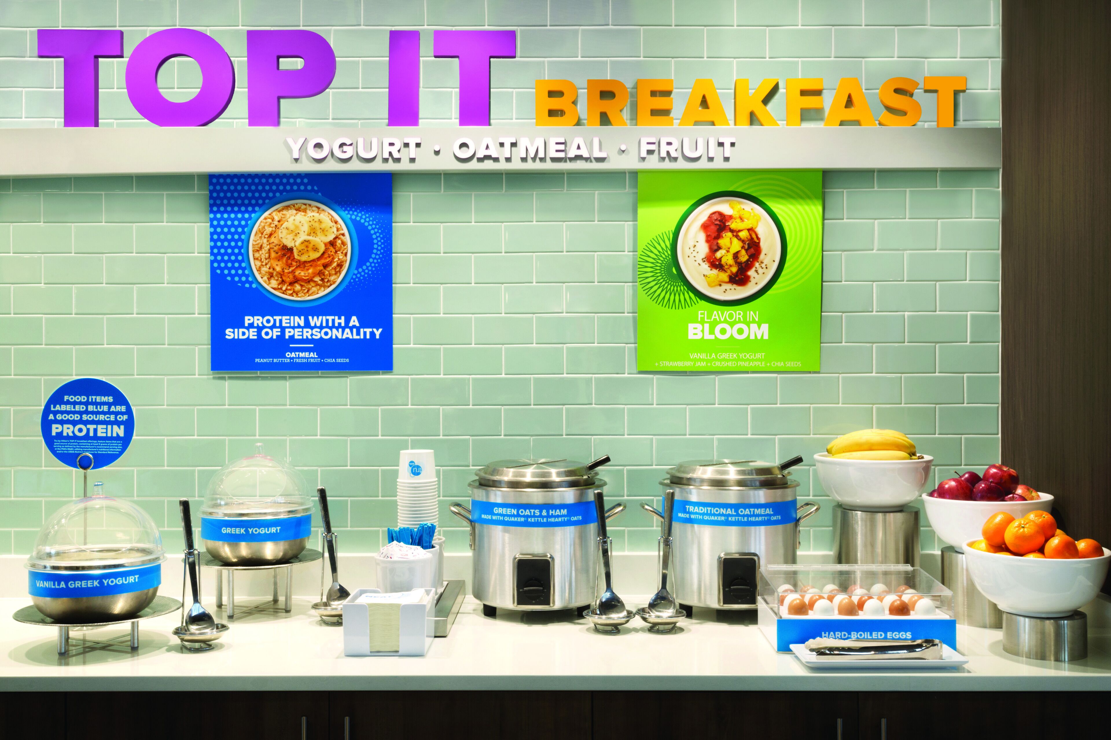 Buffet de café da manhã com alimentos identificados, incluindo iogurte, aveia e frutas.