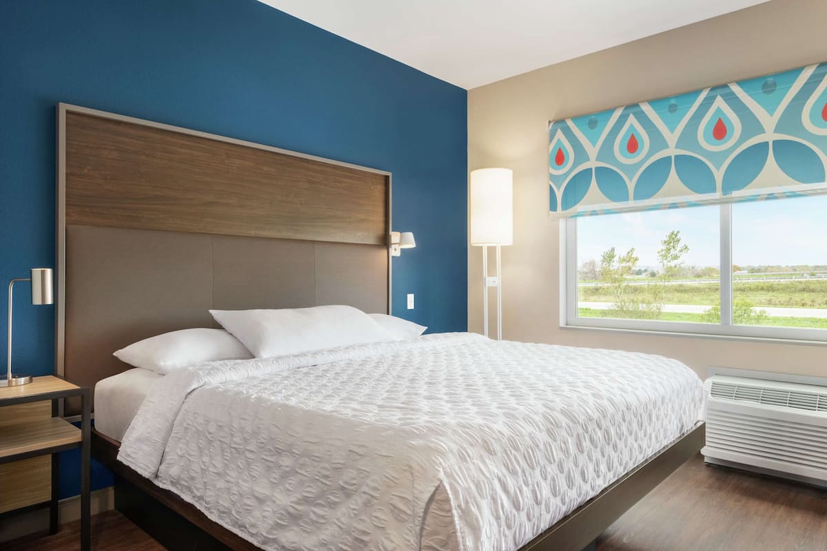 A modern hotel room with a large bed, a nightstand, a desk, a window with a view, and a decorative wall panel.