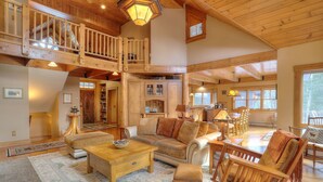 A spacious living area with a high ceiling, wooden beams, and a cozy fireplace. There is a dining area with wooden furniture and a large window providing natural light. - Potter's Place Main House - One of the Rangeley Lakes Region's finest private rental opportunities! (Rangeley Lake)