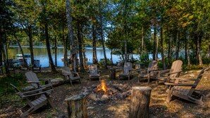 A campfire surrounded by Adirondack chairs in a forested area with a lake in the background. - Potter's Place Main House - One of the Rangeley Lakes Region's finest private rental opportunities! (Rangeley Lake)