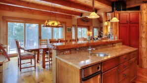 A spacious kitchen with granite countertops, wooden cabinets, and a large island with a sink. There are hanging pendant lights and a view of the outdoors through large windows. - Potter's Place Main House - One of the Rangeley Lakes Region's finest private rental opportunities! (Rangeley Lake)