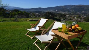 Deux fauteuils en bois sont placés sur une pelouse verdoyante, près d’une table où se trouvent des fruits et une boisson. L’ensemble offre une vue panoramique sur une ville et des montagnes.