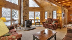A cozy cabin interior with a stone fireplace, wooden furniture, and large windows offering a view of a snowy landscape.