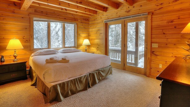 A cozy wooden cabin bedroom with a bed, two bedside lamps, a dresser, and a window with a view of snow-covered trees.