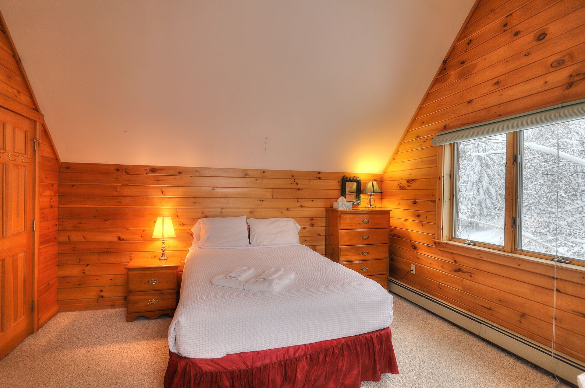 A cozy bedroom with a wooden sloped ceiling, a bed with white bedding, a nightstand with a lamp, a dresser, and a window with a snowy view.