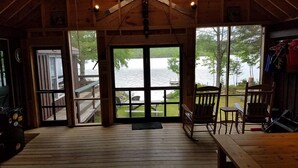 Une cabane dotée d’une terrasse en bois, d’une grande fenêtre donnant sur un lac et d’un coin repas avec des chaises et une table.