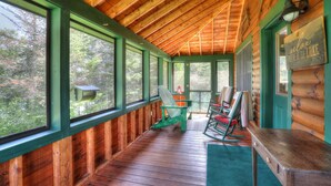 A log cabin with a wooden ceiling and large windows. There are several Adirondack chairs and a table on the porch.