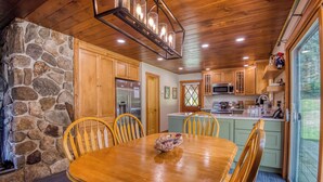 A wooden dining table with chairs, a stone wall, and a kitchen area with wooden cabinets and appliances. - 🐾23 Dolman - Amalaran - 360' of frontage on Dodge Pond, dog friendly (Rangeley)