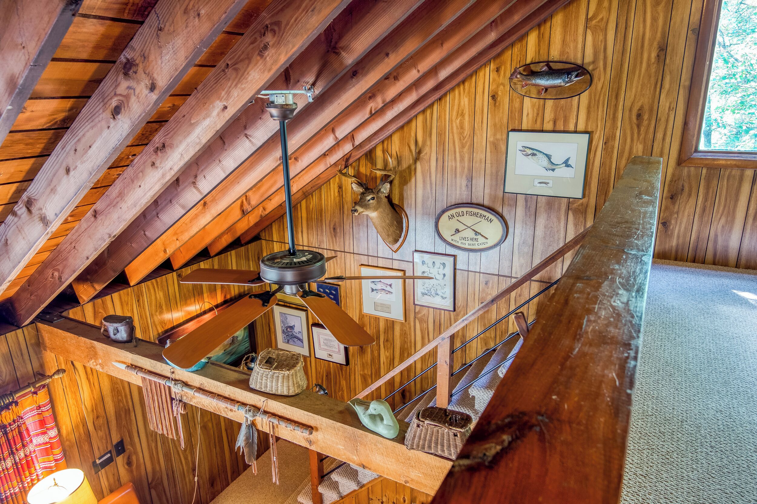 A rustic wooden interior with a slanted ceiling, wooden beams, a ceiling fan, and various fishing memorabilia on the walls.
