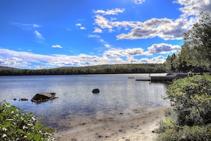 Un lago tranquilo con un muelle, rodeado de un bosque denso y un cielo azul despejado.