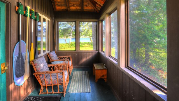 A wooden cabin with a porch, wicker chairs, a small wooden stool, and a paddle leaning against the wall.