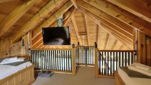 A wooden attic bedroom with a TV mounted on the ceiling, a bed with a green bedspread, and a small desk with a computer.