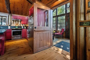 Interior de madera con cocina, comedor con una silla y un ventanal grande.