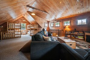 A cozy loft-style room with a high ceiling, wooden beams, a sofa, a fireplace, and a bookshelf. - Breakwater Lodge - Gorgeous home on the Eastern Shore of Mooselookmeguntic Lake (Rangeley)