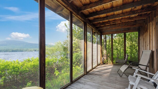 Eine Holzterrasse mit großen Fenstern mit Blick auf einen See und Wald. Zwei Liegestühle und ein kleiner Tisch sind vorhanden.