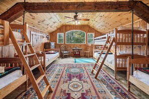 A cozy attic bedroom with wooden walls and a sloped ceiling, featuring a bed, two chairs, a small table, and a window overlooking greenery.