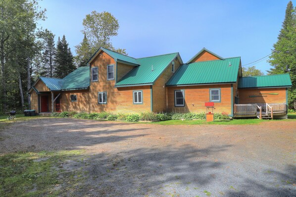 A wooden cabin with a green roof, a gravel driveway, and a fenced front yard.