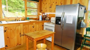 Une cuisine avec des meubles en bois, un réfrigérateur en acier inoxydable, une table en bois sur laquelle se trouve un saladier, et une fenêtre donnant sur un paysage verdoyant.