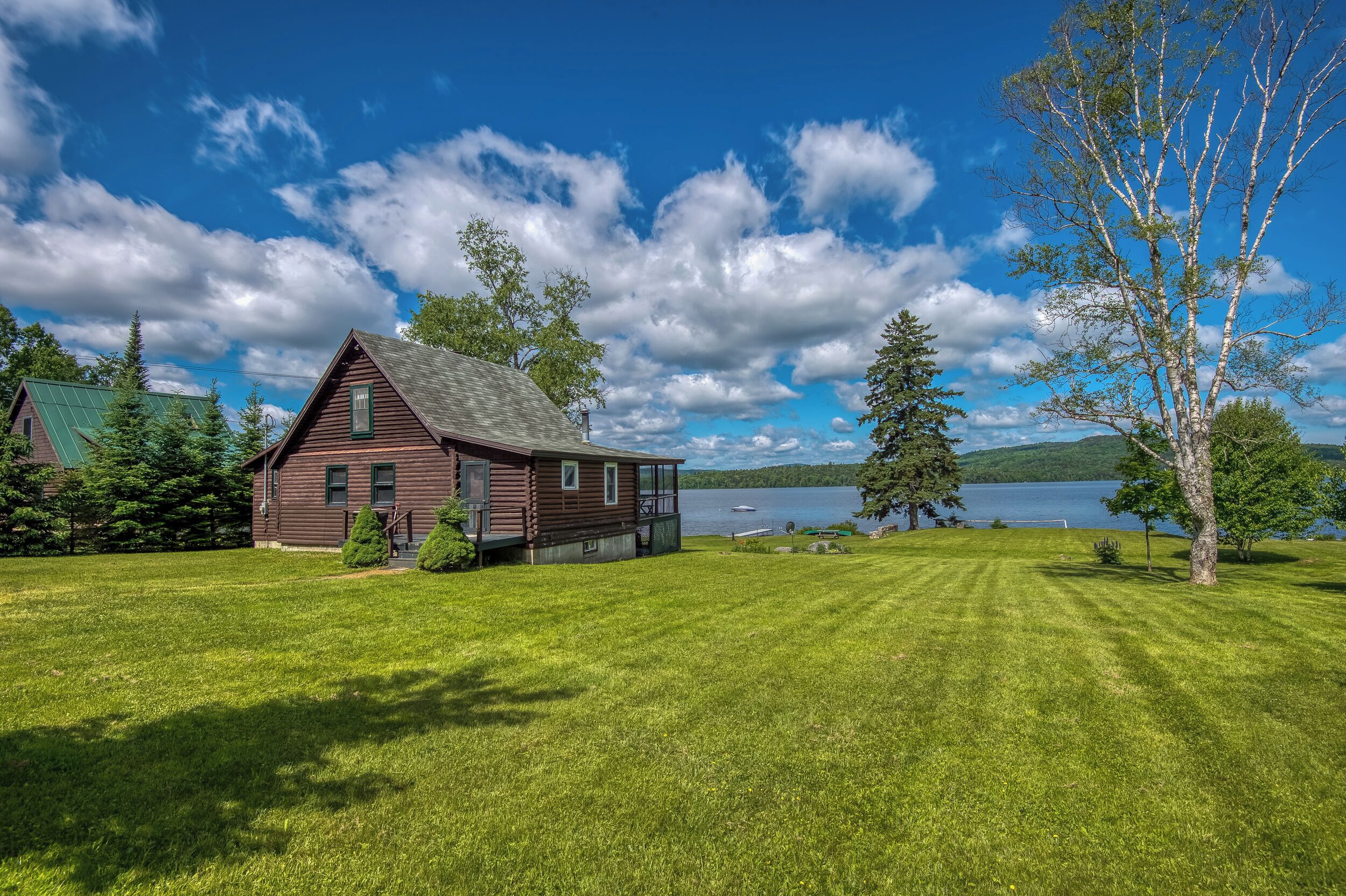 A rustic cabin with a green roof, surrounded by a well-maintained lawn and a large body of water in the background.