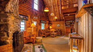 Interior acolhedor de uma cabana de madeira com lareira de pedra, móveis rústicos e um teto alto com vigas aparentes.