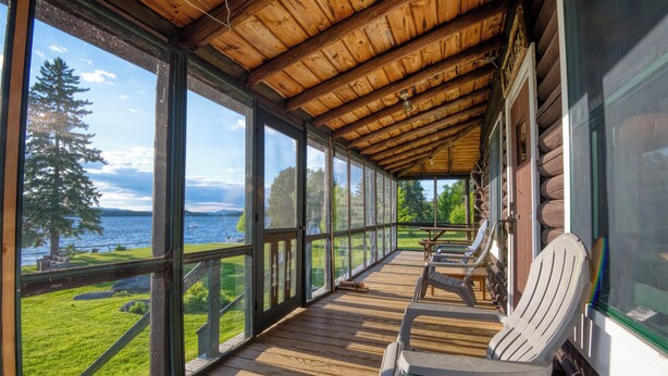Um deck de madeira com vista para um lago e floresta. Há duas cadeiras Adirondack e uma mesa.