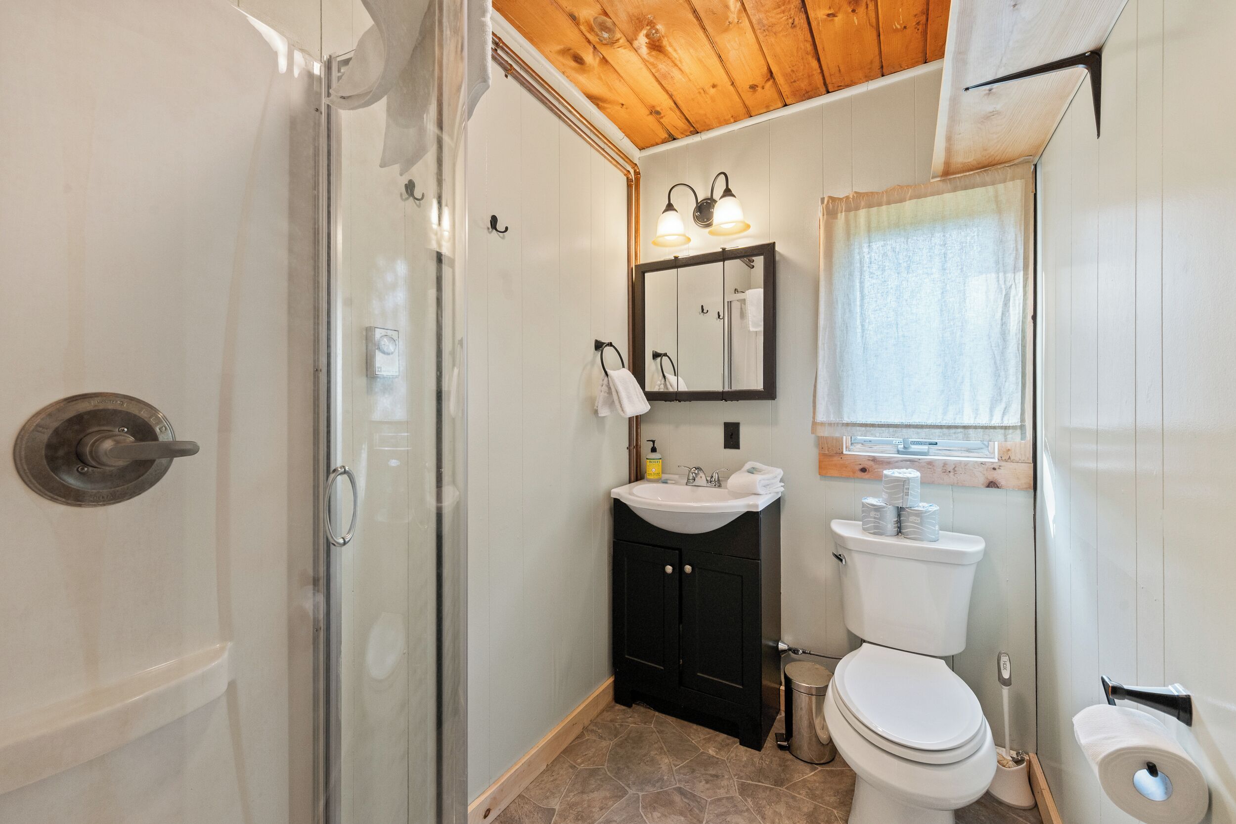 Un baño con cabina de ducha de vidrio, un lavabo con espejo, un inodoro y una ventana con techo de madera.