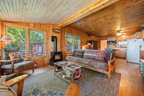 A cozy cabin interior with a fireplace, wooden furniture, and large windows. - Four Loons | Waterfront | Pet Friendly (Oquossoc)