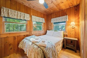 A wooden cabin bedroom with a bed, two windows with curtains, a bedside table, and a lamp.