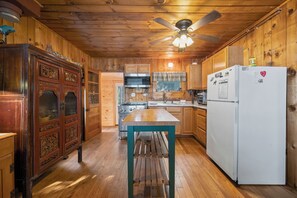 A wooden cabin interior with a kitchenette, a ceiling fan, and a wooden dining table.