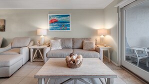 Un salon chaleureux avec un canapé, deux tables d’appoint avec des lampes, une table basse en bois avec un panier, et un tableau représentant une scène de plage.