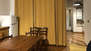 A dining area with a wooden table and chairs, a yellow curtain, a white refrigerator, and a kitchen visible in the background. - Macci Number Eight (Florence)