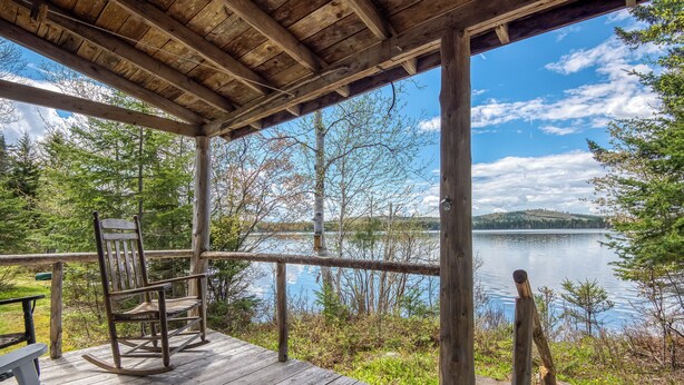 A wooden deck with a rocking chair and a bench overlooking a lake, surrounded by trees and a clear sky. - 🐾Gray Wulff - This rustic dog friendly camp sits on the shore of Quimby Pond (Rangeley)
