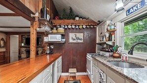 Une cuisine compacte avec des meubles en bois, un four à micro-ondes, une cuisinière et un évier. Il y a une fenêtre donnant sur des arbres et un panneau indiquant « LAC ».