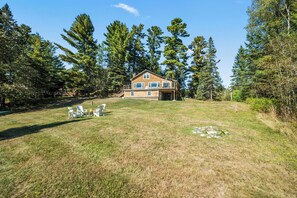 A two-story wooden house with a deck, surrounded by a grassy yard and tall trees.