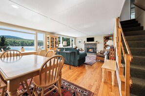 A spacious living room with a wooden dining table, chairs, a sofa, and a fireplace. There is a staircase on the right and a large window offering a view of the outdoors.