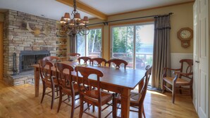 A dining area with a wooden table and chairs, a stone fireplace, and a view of the outdoors through large windows. - Beach Front Retreat - Private home on Mooselookmeguntic Lake (Oquossoc)