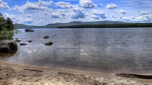A calm lake with a boat, rocky shoreline, and distant mountains under a partly cloudy sky. - Beach Front Retreat - Private home on Mooselookmeguntic Lake (Oquossoc)