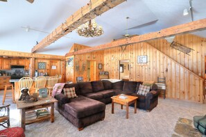A rustic cabin interior with a wooden dining area, a large sofa, and a staircase leading to a loft. - Saddleback Lake Lodge 25 | Close to Saddleback (Rangeley)