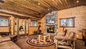 A log cabin interior with a stone fireplace, wooden furniture, and a large window overlooking a forest. - 🌲 Bird's Perch View 🌅 Custom Cabin, Mountain Views, Hot Tub & Fire Pit (Sevierville)