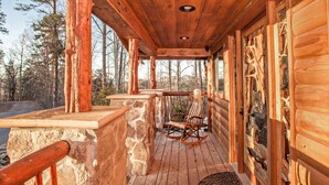 A wooden deck with a stone wall, a rocking chair, and a rug. - 🌲 Bird's Perch View 🌅 Custom Cabin, Mountain Views, Hot Tub & Fire Pit (Sevierville)