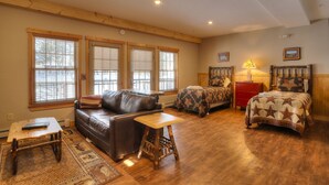 A cozy room with a wooden floor, two beds with patterned quilts, a leather sofa, a small wooden table, and a red nightstand. - Oquossoc Suite #6 | A/C & Fireplace (Oquossoc)
