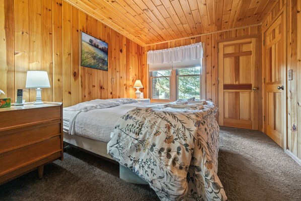 A wooden-paneled bedroom with a bed, a dresser, a window with a view, and a door. - Saddleback Lake Lodge 19 | Close to Saddleback (Rangeley)