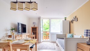 A modern, well-lit living room with a dining area, a television, and a large window offering a view of greenery. - Casa Montana II- Country Apartment (Balatonalmadi)