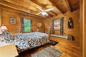 A wooden cabin bedroom with a bed, two windows, a ceiling fan, and a small shelf with stuffed animals.