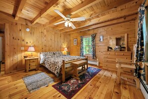 Interior de una cabaña de madera con una cama, un escritorio, una silla, un ventilador de techo y una ventana con cortinas.