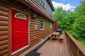 Una cabaña de troncos con puerta roja, una terraza y una parrilla.