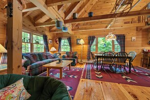 A cozy cabin interior with a wooden ceiling, a sofa, a dining table with chairs, and a patterned rug.