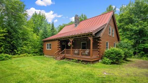 A log cabin with a porch and a chimney, surrounded by lush greenery.